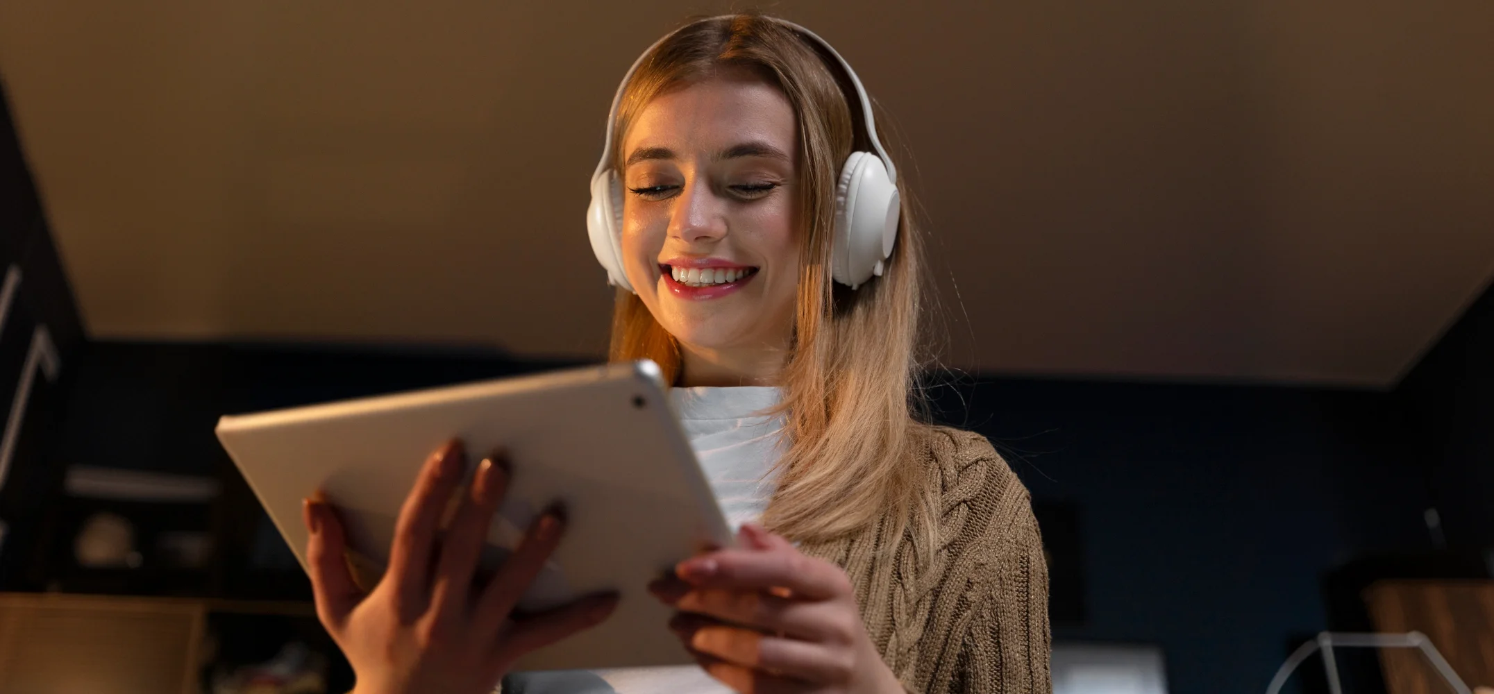 A woman in headphones smiling at a tablet she holds in both hands